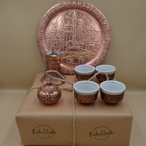 Può includere: Un set da caffè in rame composto da un vassoio rotondo sbalzato con una scena urbana, una caffettiera, un piccolo contenitore con coperchio e quattro tazze in ceramica bianca con supporti in rame. Gli oggetti sono disposti su una scatola con la scritta Rahatlook.