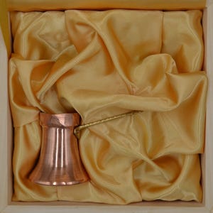 Puede incluir: Una cafetera de cobre pulido con base ancha, cuello estrecho y un mango largo de latón texturizado. La cafetera está colocada sobre un forro de tela satinada de color dorado claro dentro de una caja de madera cuadrada, creando un aspecto metálico.