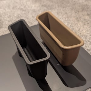 Puede incluir: Dos moldes rectangulares para hornear, uno negro y otro beige, sobre una bandeja metálica gris. Los moldes están vacíos y tienen forma de molde para pan. La bandeja tiene esquinas redondeadas y agujeros.