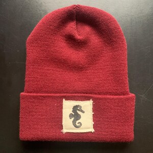 May include: A burgundy knit beanie with a folded brim. A small, off-white square patch is sewn on the front, featuring a black seahorse design. The hat is made of a soft, textured material.