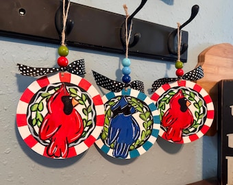 May include: Three circular wooden ornaments hanging from a black rack. Two ornaments feature a red cardinal bird, and one features a blue bird, each centered within a green leaf wreath. They have black and white polka dot bows and colorful beads.