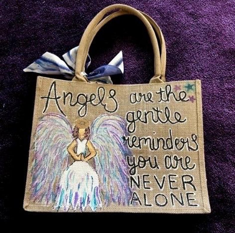 Peut inclure: Un sac fourre-tout en toile de jute beige avec des poign&eacute;es marron et un n&oelig;ud bleu et blanc. Le sac pr&eacute;sente un ange peint &agrave; la main avec des ailes violettes et bleues et le texte "Angels are the gentle reminders you are NEVER ALONE."