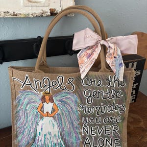 Peut inclure: Un sac fourre-tout en toile de jute beige avec des poign&eacute;es marron. Le sac pr&eacute;sente un ange peint &agrave; la main avec des ailes color&eacute;es et le texte "Angels are the gentle reminders you are NEVER ALONE." Un foulard rose est nou&eacute; &agrave; la poign&eacute;e.