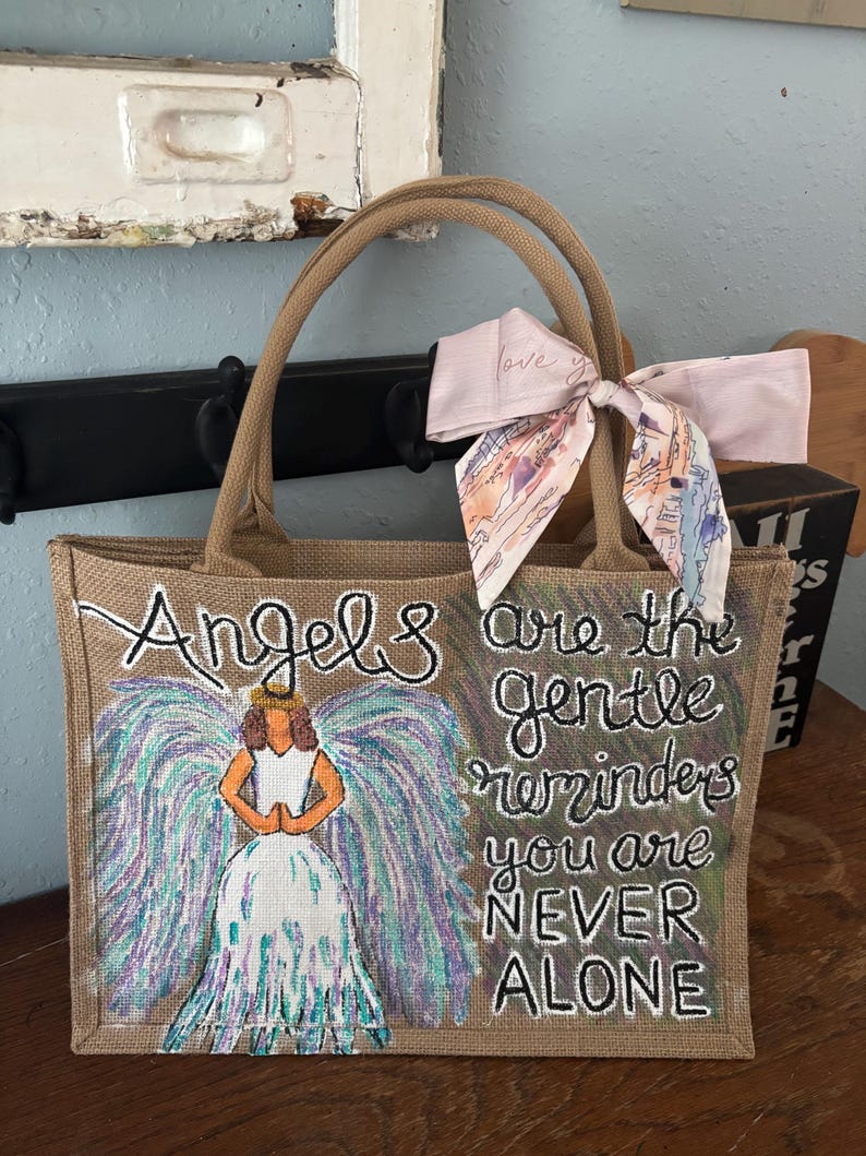 Peut inclure: Un sac fourre-tout en toile de jute beige avec des poign&eacute;es marron. Le sac pr&eacute;sente un ange peint &agrave; la main avec des ailes color&eacute;es et le texte "Angels are the gentle reminders you are NEVER ALONE." Un foulard rose est nou&eacute; &agrave; la poign&eacute;e.