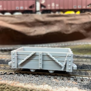 May include: A light gray model train freight car with diagonal bracing and rivets. The miniature car sits on a model railroad track with a brown and green landscape in the background. The car has four wheels.