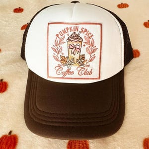 Puede incluir: Gorra de camionero marrón y blanca con la parte trasera de malla. El panel frontal presenta un diseño bordado con las palabras "Pumpkin Spice Coffee Club" y una ilustración de un café con leche de calabaza especiada.