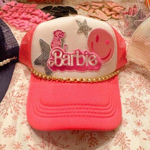 May include: A pink trucker hat with a white front panel featuring the word "Barbie" in pink script. The hat is decorated with a smiley face, stars, and a small pink dog. A gold chain accents the brim.