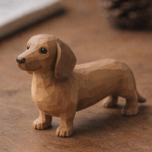 Könnte beinhalten: Eine kleine, handgeschnitzte Holzfigur eines Dackels. Die Skulptur zeigt eine hellbraune Holzmaserung mit sichtbaren Meißelspuren am ganzen Körper, eine glatte Schnauze, dunkle Augen und Schlappohren in einer klassischen, langgestreckten Pose.