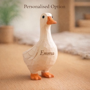 May include: A carved wooden duck figurine with a light beige finish, an orange beak, and orange feet. The text "Personalised Option" appears above the duck, and the name "Emma" is engraved on the chest of the duck, which has visible wood grain texture.