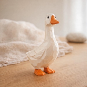 May include: A hand-carved wooden figurine of a goose with a smooth, off-white body and a bright orange beak and feet. The sculpture features visible faceted carving marks across its surface, a small black eye, and a long, upright neck.