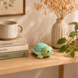 May include: A small, carved turtle figurine with a light green shell and a smiling face with pink cheeks. The turtle has a textured, faceted surface and is positioned on a wooden surface next to a stack of three books and a ceramic mug.