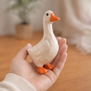 May include: A small, hand-carved wooden figurine of a goose with a smooth, off-white body and a bright orange beak and feet. The surface features visible, faceted carving marks throughout, giving the bird a textured, geometric appearance.