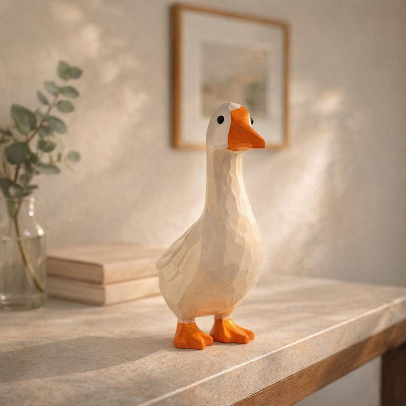May include: A carved wooden goose figurine featuring a smooth, off-white body with visible chisel marks, a bright orange beak, and orange webbed feet. The goose has small, round black eyes and is standing in an upright, alert posture.