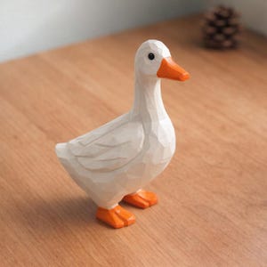 May include: A white goose figurine with a faceted, geometric surface texture. The bird features a bright orange beak and matching orange webbed feet. It has a small black eye and is standing in a profile position, showcasing its angular, carved design.