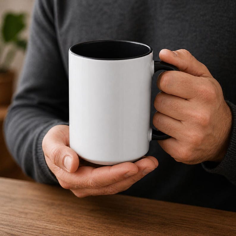 May include: A white ceramic mug with a black interior and handle. The mug is held by a person, showcasing its clean design. The mug is a standard size, perfect for coffee or tea.