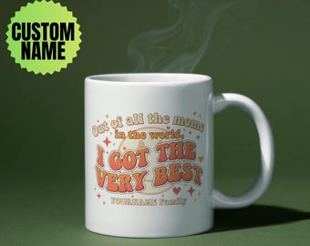 May include: White ceramic mug with the text "Out of all the moms in the world, I GOT THE VERY BEST" in orange and cream colors. The mug also has the text "YOURNAME Family" and a green custom name tag.