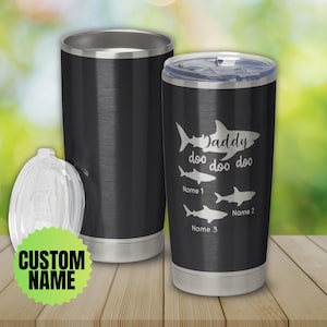 May include: Black stainless steel tumbler with a lid, featuring a shark design and the words "Daddy doo doo doo" with space for custom names. A clear lid and a blank tumbler are also visible.