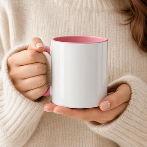 May include: A white ceramic mug with a pink interior and handle. The mug is held in both hands, with the person wearing a cream-colored sweater. The mug is a standard size for coffee or tea.