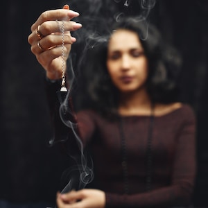 May include: A silver pendulum with a triangular crystal is held in front of a woman. Smoke swirls around the pendulum and the woman, who is wearing a maroon top and a necklace. A lit candle is in the background.