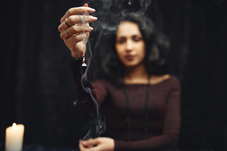 May include: A silver pendulum with a triangular crystal is held in front of a woman. Smoke swirls around the pendulum and the woman, who is wearing a maroon top and a necklace. A lit candle is in the background.
