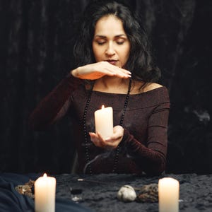 May include: A person in a dark burgundy off-the-shoulder top holds a lit white candle, with two other candles and a black beaded necklace. The scene is set on a black table with a dark background, evoking a mystical theme.