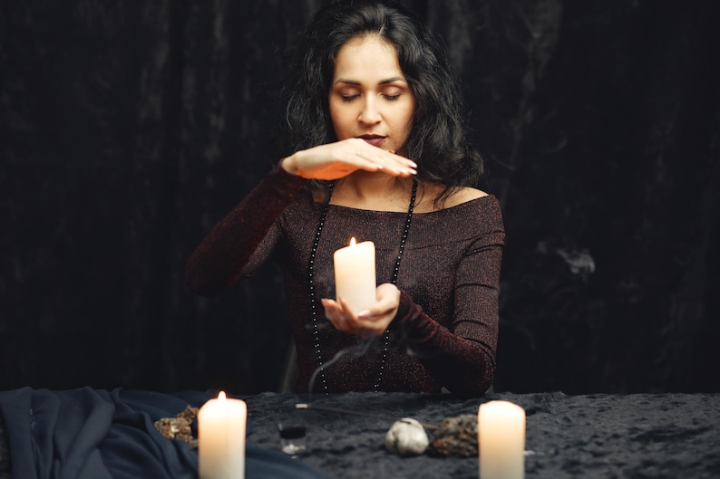 May include: A person in a dark burgundy off-the-shoulder top holds a lit white candle, with two other candles and a black beaded necklace. The scene is set on a black table with a dark background, evoking a mystical theme.