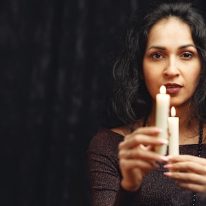 May include: A woman holding two lit candles against a dark background. The candles are white and have a soft glow. The woman is wearing a dark, off-the-shoulder top and has long, dark hair.