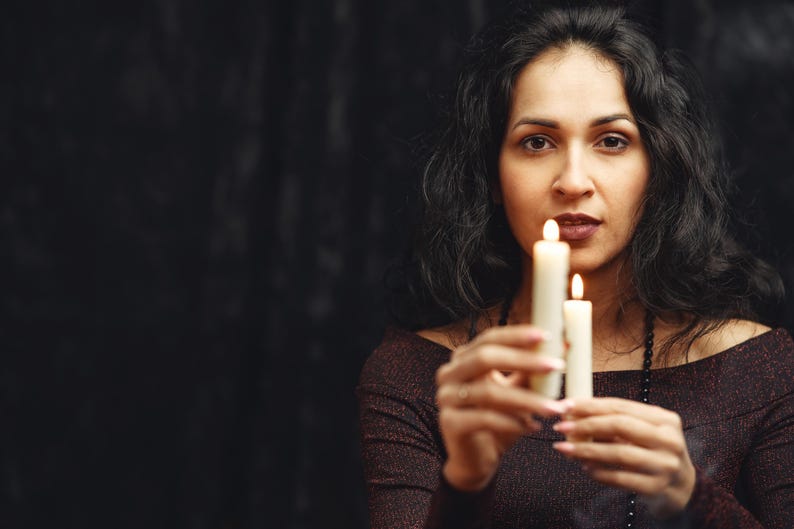 May include: A woman holding two lit candles against a dark background. The candles are white and have a soft glow. The woman is wearing a dark, off-the-shoulder top and has long, dark hair.