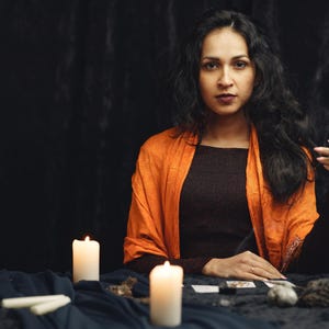 May include: A woman with long, dark hair wearing a brown top and orange shawl sits at a table. Lit candles and tarot cards are on the table. She holds a string of beads, creating a mystical atmosphere.