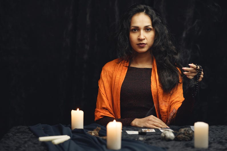 May include: A woman with long, dark hair wearing a brown top and orange shawl sits at a table. Lit candles and tarot cards are on the table. She holds a string of beads, creating a mystical atmosphere.