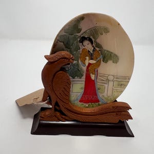 May include: A carved wooden sculpture featuring a phoenix and a painted scene of a woman in traditional attire. The artwork is mounted on a dark brown base, showcasing intricate details and vibrant colors.