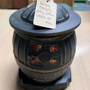 May include: A black McCoy Pottery Belly Stove cookie jar with a lid. The jar has a stove-like design with decorative floral accents in red, yellow, and orange. The tag reads "McCoy Pottery Belly Stove cookie Jar".