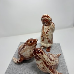 May include: Ceramic figurines of seals and a person in a winter coat. The seals are in a lying down position, with a marbled brown and white design. The person is standing, also with a marbled brown and white design. The figurines are on a gray surface.