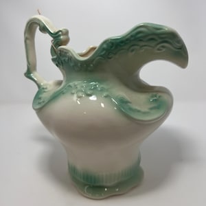 May include: A vintage ceramic pitcher with a cream-colored body and seafoam green accents. The pitcher features a decorative handle and a scalloped rim with embossed floral details. The base and rim are also accented with the same green color.