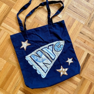 May include: Navy blue tote bag with long handles. The bag features a white and blue applique with the letters "NYC" and three star-shaped patches. The bag is made of a sturdy fabric and is suitable for carrying various items.