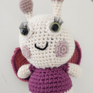 May include: A handmade crochet butterfly toy with a white body, purple dress, and red and purple wings. The butterfly has large, googly eyes with long eyelashes, pink cheeks, and a smiling mouth. The antennae are cream-colored with purple tips.
