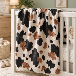 May include: A soft, plush blanket featuring a tri-color cow print pattern with irregular patches of black, brown, and cream. The blanket is draped over the side of a white wooden crib, showcasing its textured, fuzzy surface and repeating animal-inspired design.