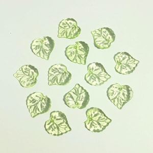 May include: A collection of twelve small, translucent light green leaf-shaped beads. Each bead features a molded, raised vein pattern on the surface, creating a textured appearance. The leaves are arranged in a scattered, circular formation.