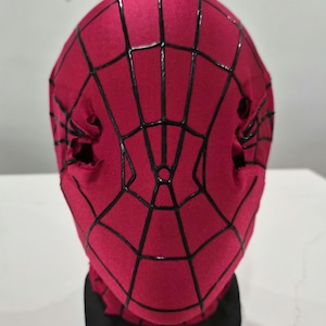 Op de afbeelding: Een rood Spider-Man-masker met een zwart webpatroon. Het masker is gemaakt van een zacht materiaal en heeft oogopeningen. Het masker wordt op een zwarte standaard getoond.