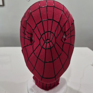 Op de afbeelding: Een rood Spider-Man-masker met zwarte webdetails. Het masker is gemaakt van stof en heeft oogopeningen. Het masker wordt gepresenteerd op een witte schuimbasis.