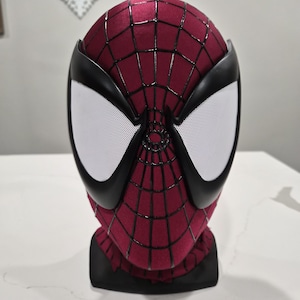 May include: A Spider-Man mask with a burgundy and black color scheme. The mask features a web pattern and large white eye lenses with black borders. The mask is designed to fit the head and face.