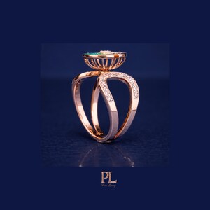 May include: Rose gold ring with a unique design. The ring features a central emerald-colored stone and is accented with small diamonds. The band has a double-loop design, with diamonds set along the curves. The ring is set against a dark blue background.