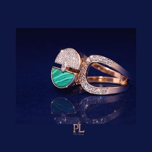 May include: A gold ring with a unique design, featuring a half-circle of sparkling diamonds and a section of green malachite. The ring has a split band and is set against a dark blue background.