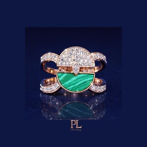 May include: Rose gold ring featuring a half-circle of sparkling diamonds and a half-circle of green malachite. The ring has a unique design with diamond accents on the band. The ring is displayed on a dark blue background.