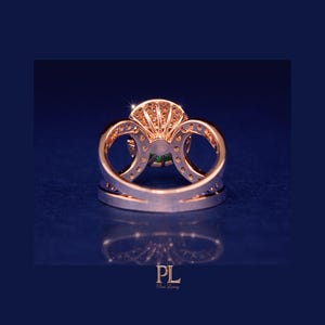 May include: A rose gold ring with a large, round green gemstone at the center, surrounded by smaller clear stones. The ring has a double band design with intricate detailing and is set against a dark blue background.