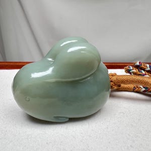 Puede incluir: Escultura de jade verde claro de un conejo. El conejo est&aacute; en posici&oacute;n enroscada con las orejas dobladas hacia atr&aacute;s. La escultura tiene una superficie lisa y pulida y est&aacute; sobre un fondo neutro. Un cord&oacute;n decorativo es visible en el fondo.