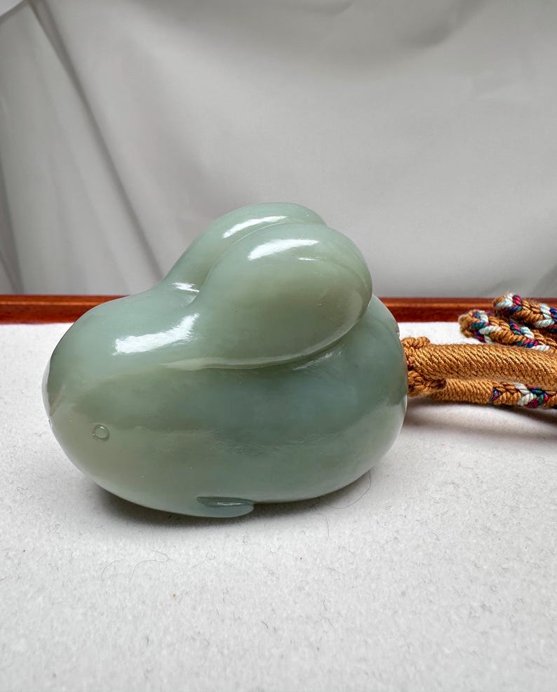 Puede incluir: Escultura de jade verde claro de un conejo. El conejo est&aacute; en posici&oacute;n enroscada con las orejas dobladas hacia atr&aacute;s. La escultura tiene una superficie lisa y pulida y est&aacute; sobre un fondo neutro. Un cord&oacute;n decorativo es visible en el fondo.