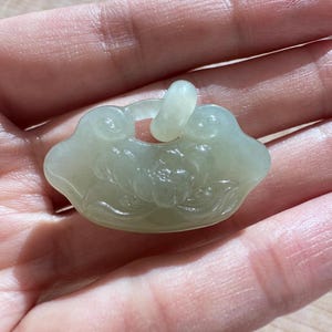 May include: A pale green, translucent jade pendant shaped like a traditional lock, featuring a carved lotus flower design on the front surface. The pendant includes a small, rounded loop at the top for attachment, showcasing a smooth, polished finish.