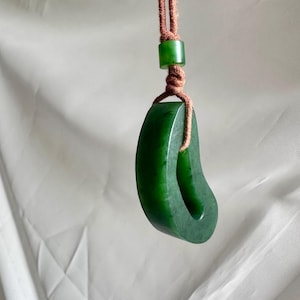 May include: A dark green, smooth, crescent-shaped stone pendant suspended from a brown cord. The pendant features a central opening and is secured by a matching green cylindrical bead above the knot, creating a simple, cohesive design.