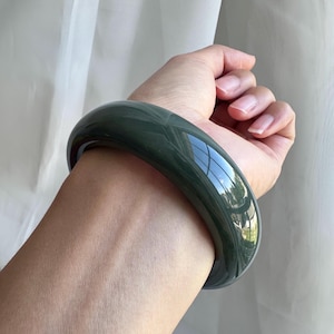 May include: A smooth, solid dark green bangle bracelet with a polished, reflective surface. The circular accessory features a thick, rounded profile and a deep, uniform forest green color that catches the light, creating a bright, white reflection.
